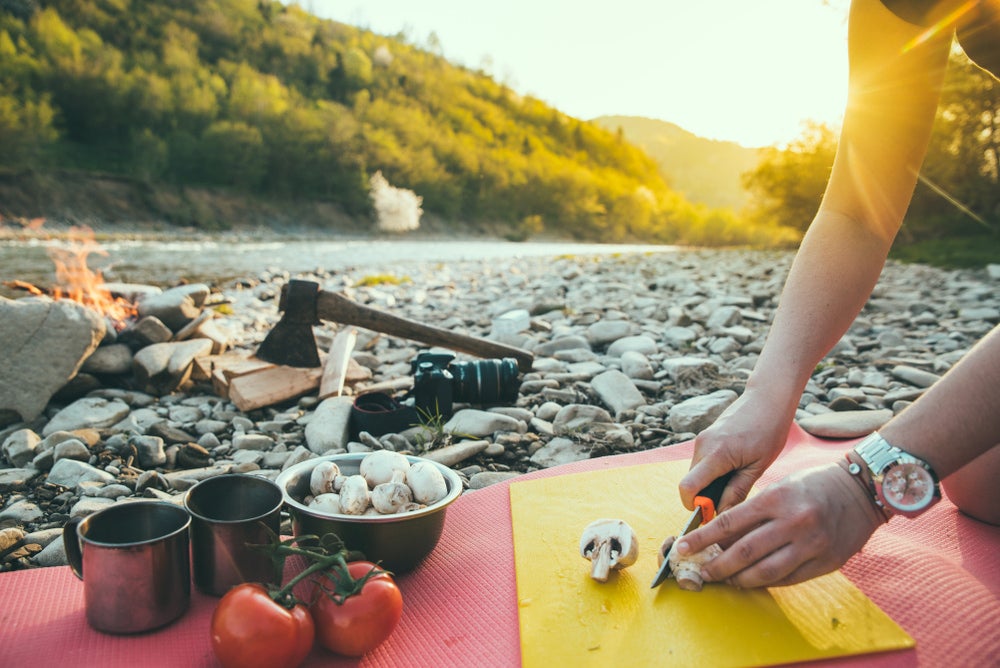 a woman slicing vegetables on a cutting board next to a campfire