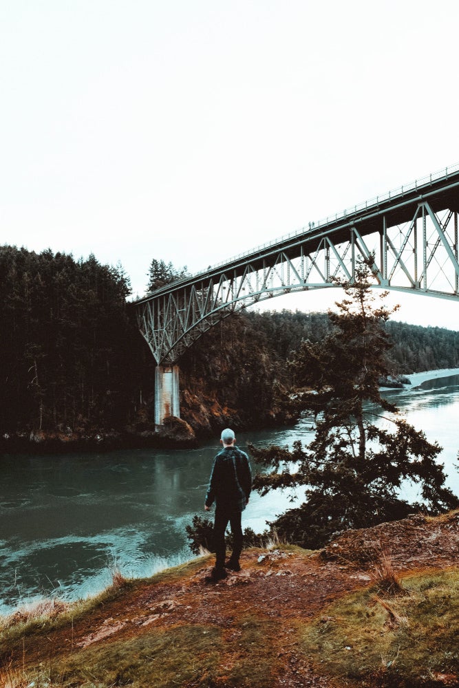 Man standing beside Pass Lake under an iron bridge.