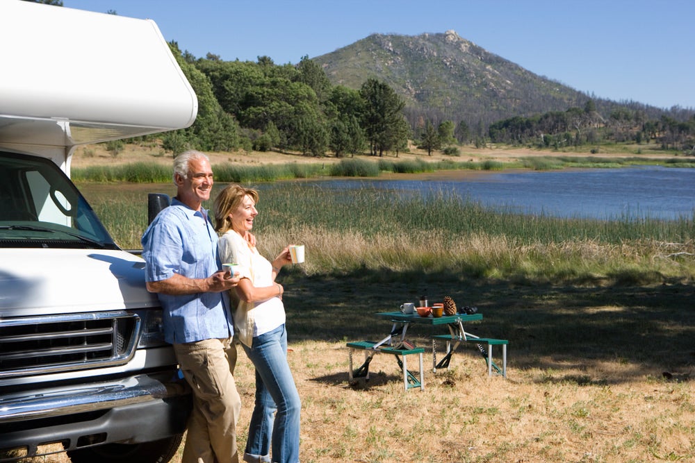 an elderly couple smiling next to a lake and leaning on their RV camper