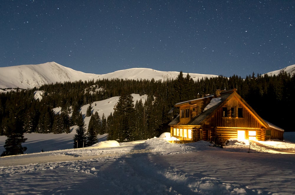 10th Mountain Division Hut lit up at night against snow covered mountains.