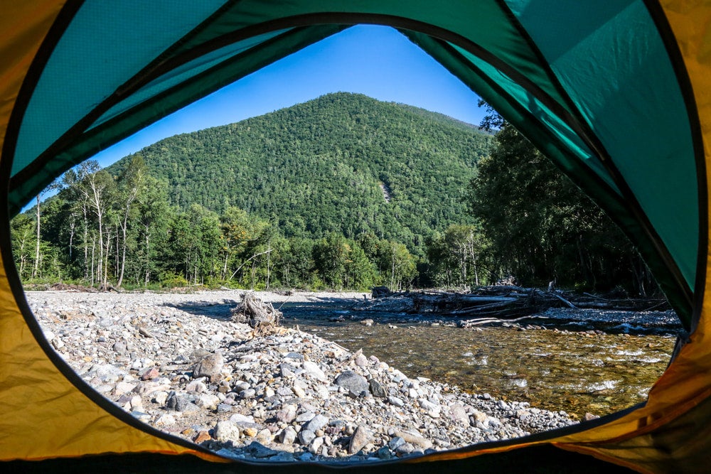 a green mountain, rocky riverbed and shallow river as seen through the door of a green tent