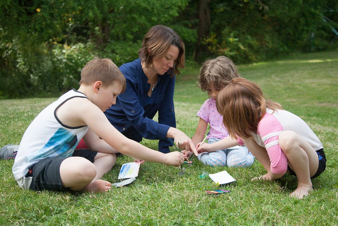 Teacher and students looking at notebooks on a grassy lawn.