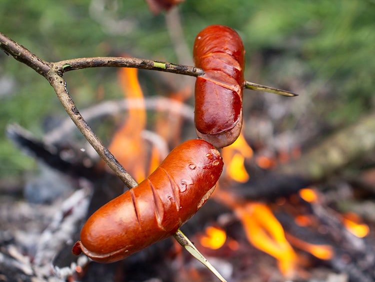 2 sliced breakfast sausages on a forked stick over a campfire