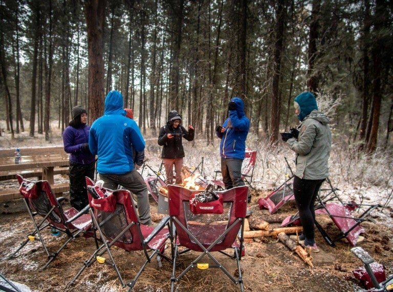 a group of campers in jackets around a campfire with camping chairs in a circle