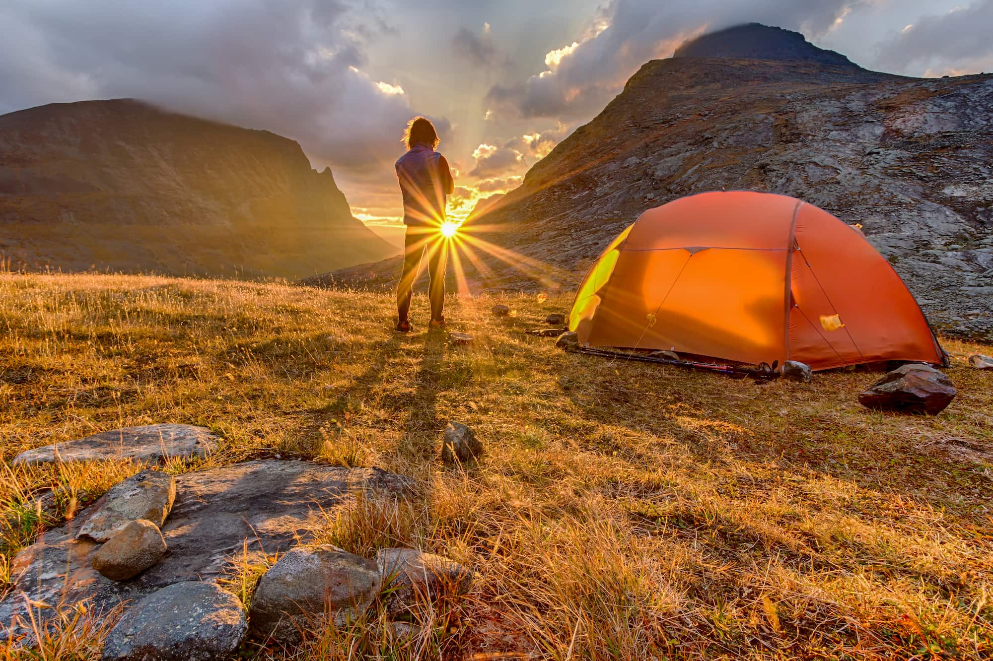 Person beside tent watching the sunrise in a mountain landscape.