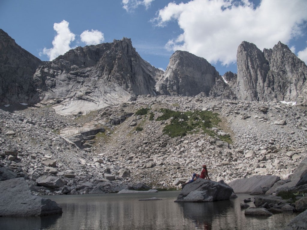 a man sits ona . rock in a lake beneath grey mountains in backcountry wyoming