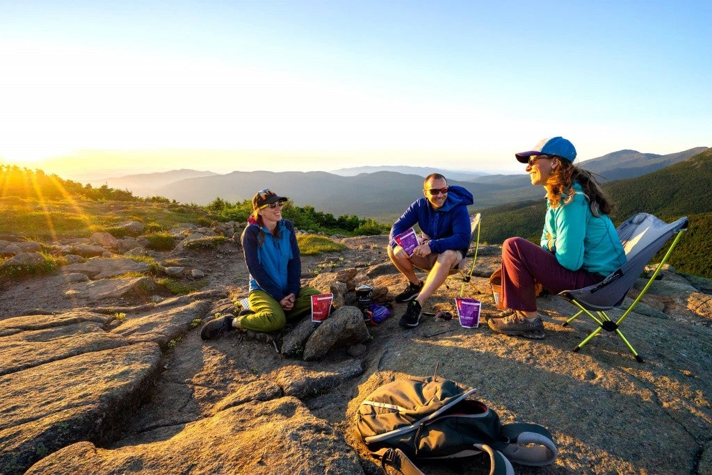 three people sitting on a rock face eating goot to go camping meals