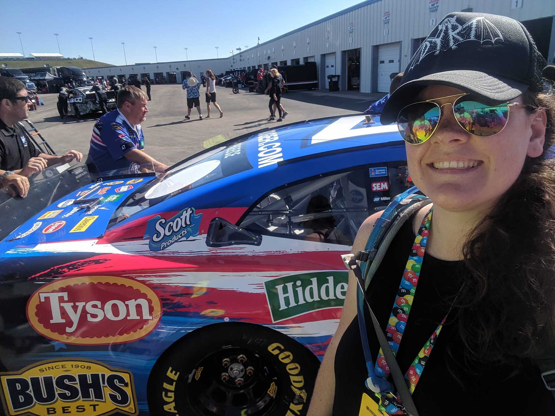 Writer with sunglasses and black baseball cap poses in front of Nascar car. 