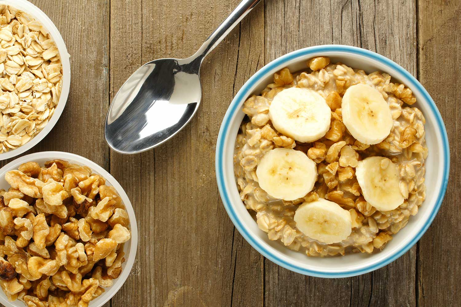Several bowls filled with granola and banana sit on a wooden table next to a spoon 