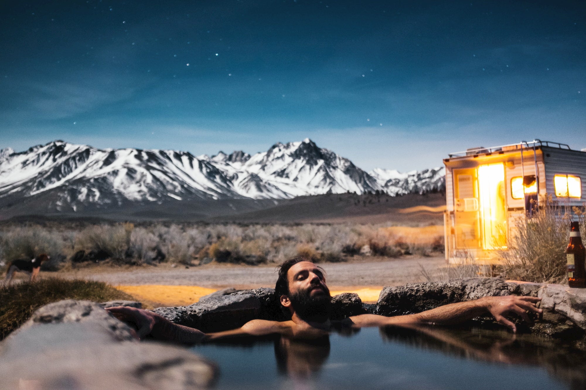 Bearded man lounging in hot springs beside camper in a mountain landscape.
