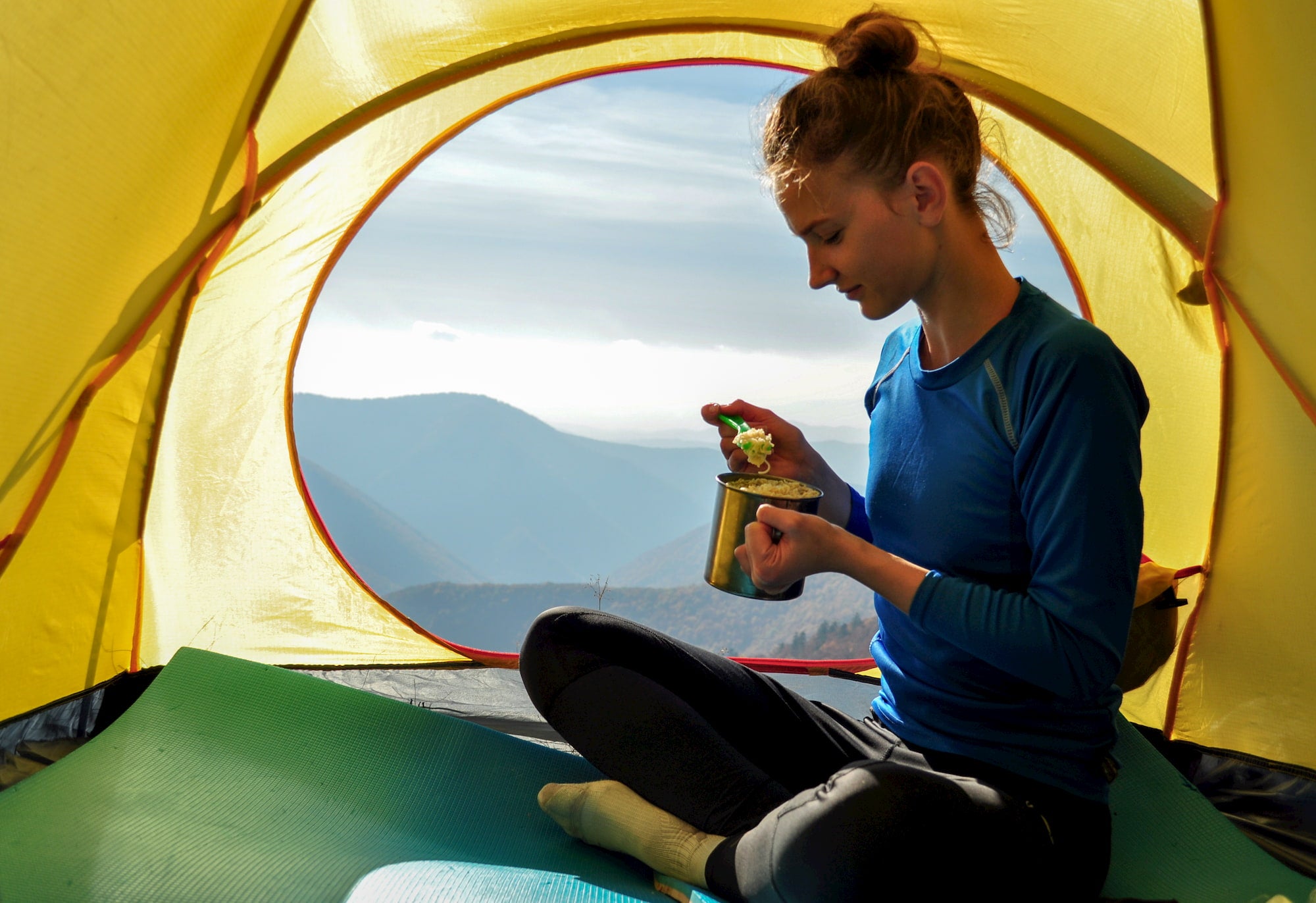 Female hiker eating food from a mug in her tent.