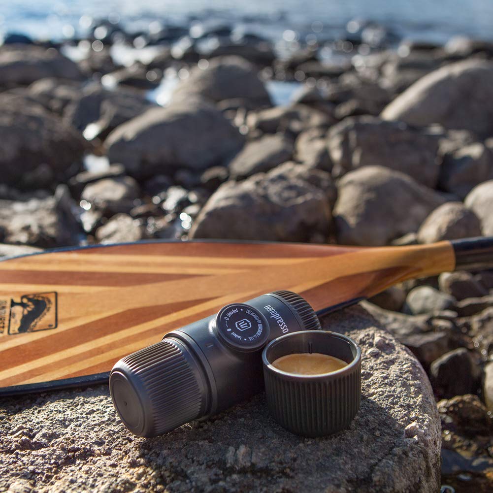 Cup of coffee and black coffee maker beside a wooden paddle on rocks by a seashore.