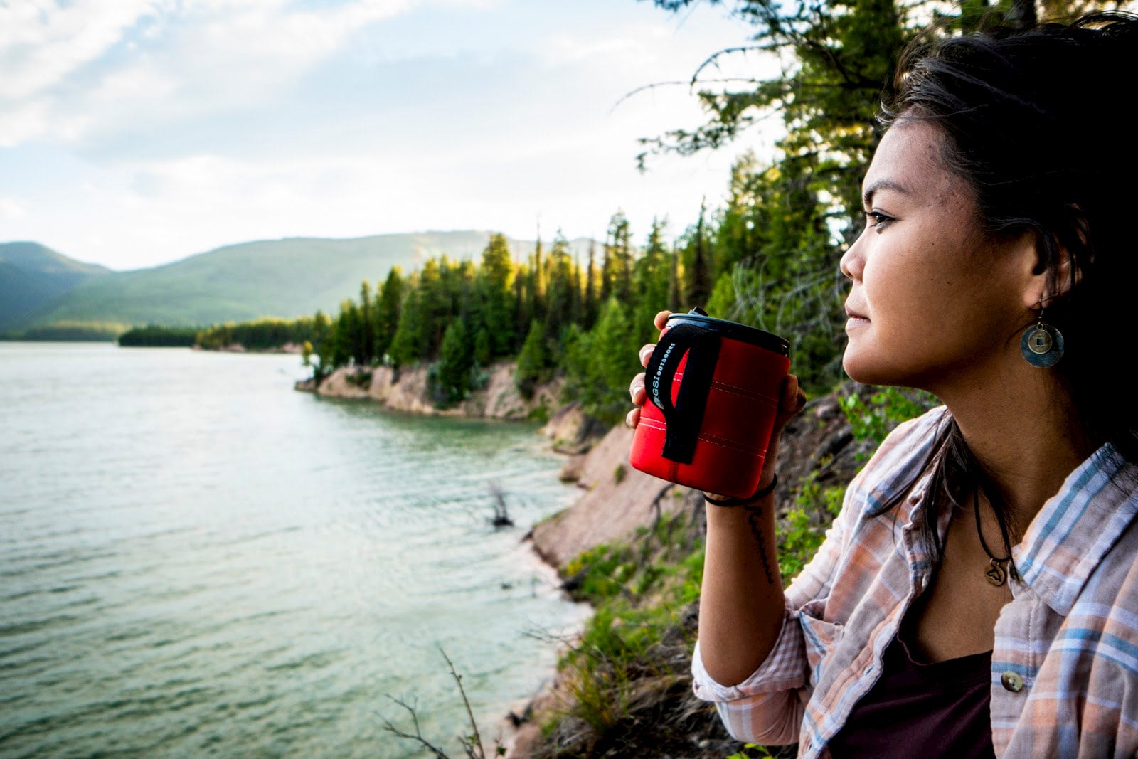 Women drinking out of red mug on the side of a lake.