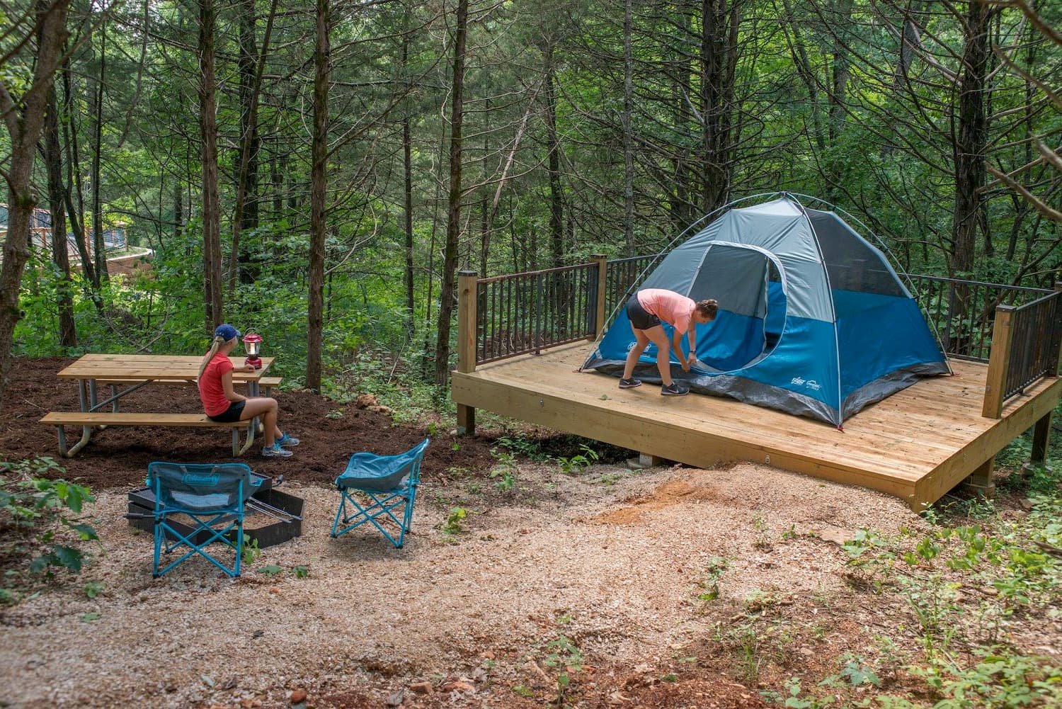 two people at campsite, one sitting on a picnic bench while the other sets up a tent