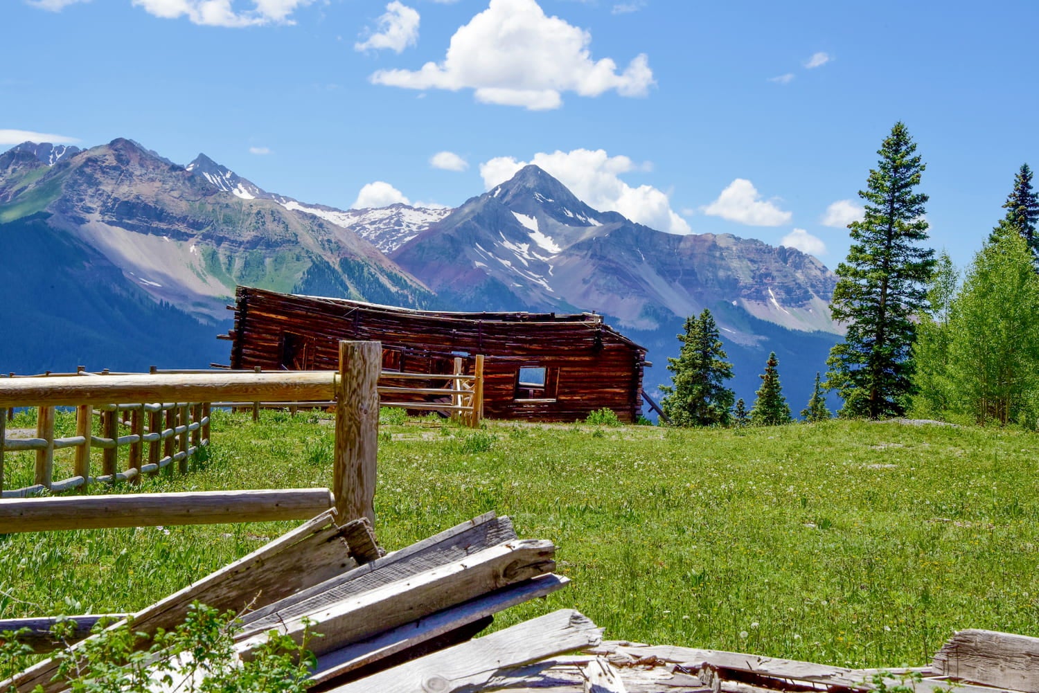 green grass, wood fence, abandoned cabin and mountain peaks in the background