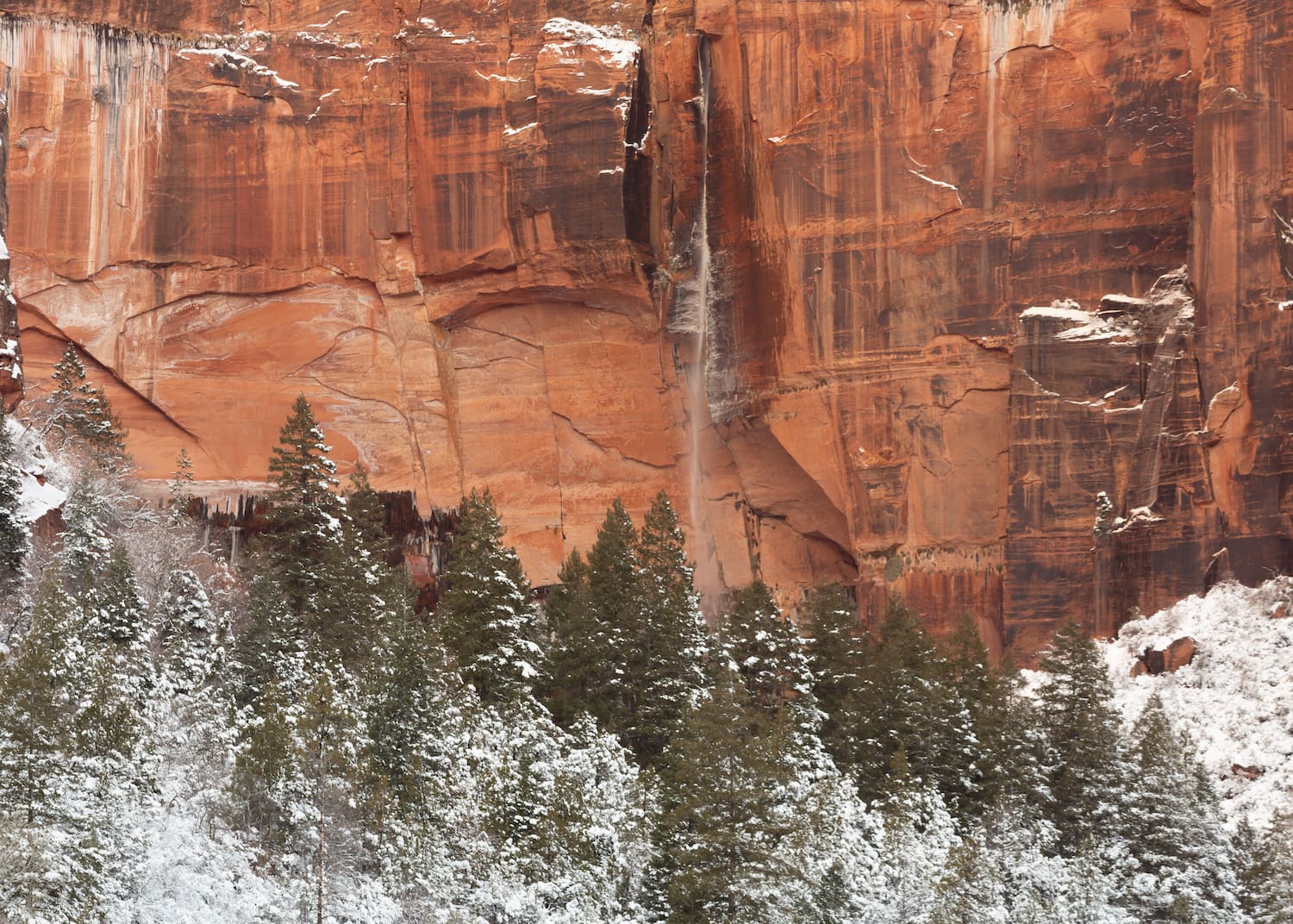 A narrow waterfall streams down a red sandstone cliff face in front of snow covered hills and pine trees.