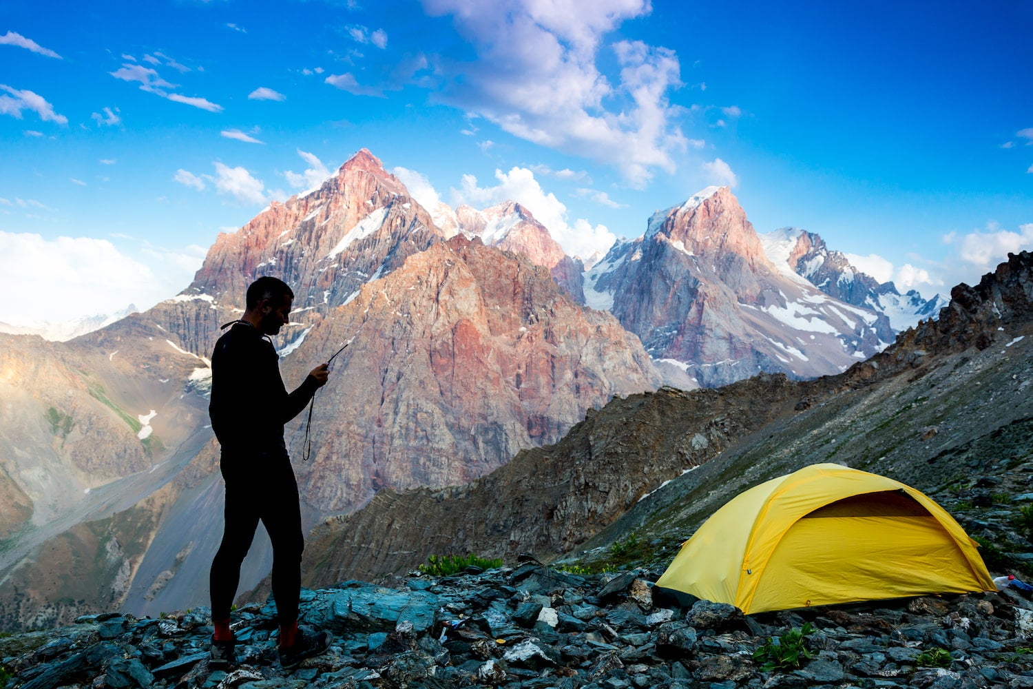man on mountain top looking at phone with tent nearby