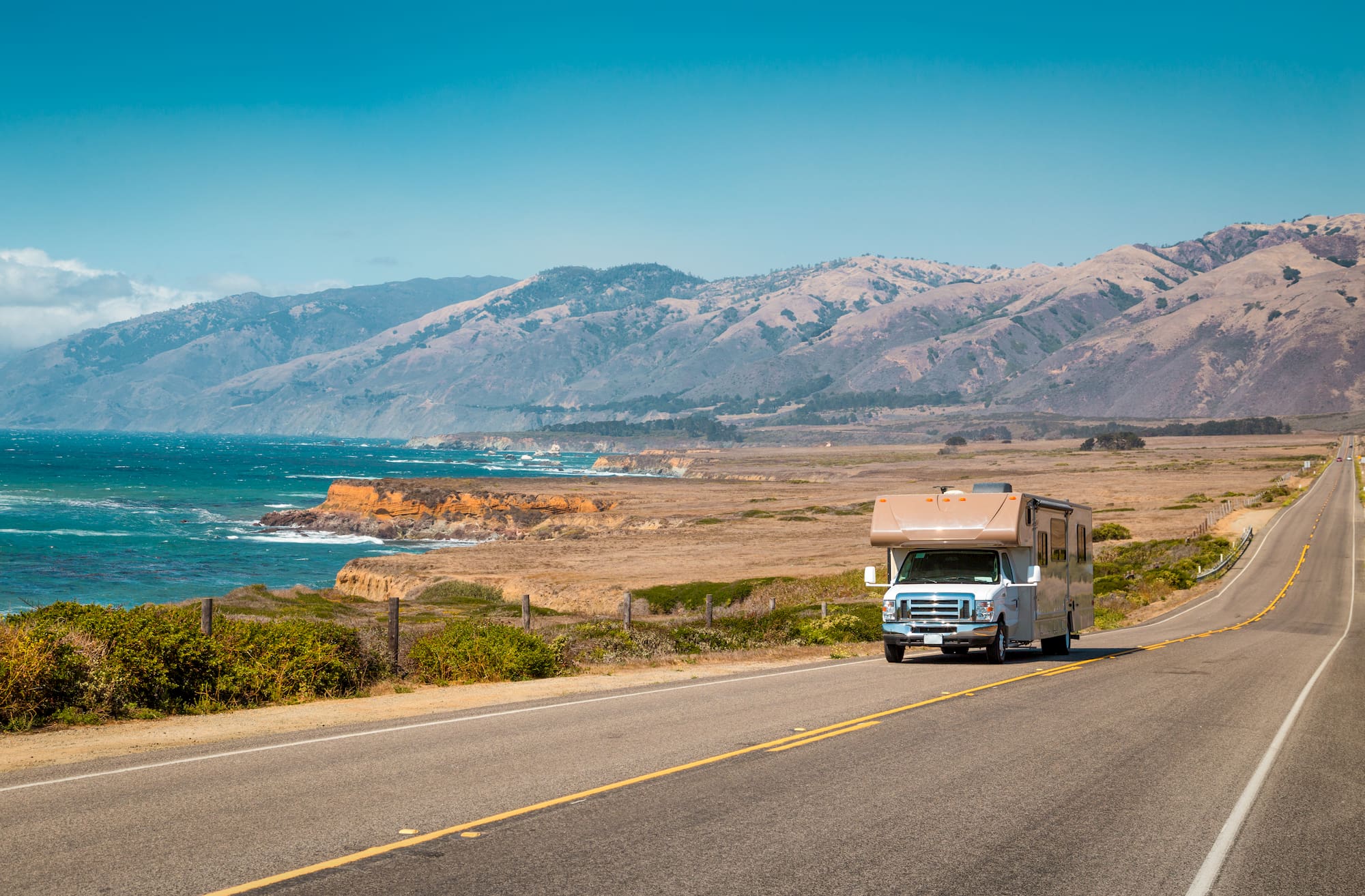 RV driving along the California coast beside the pacific ocean with mountains in the background.