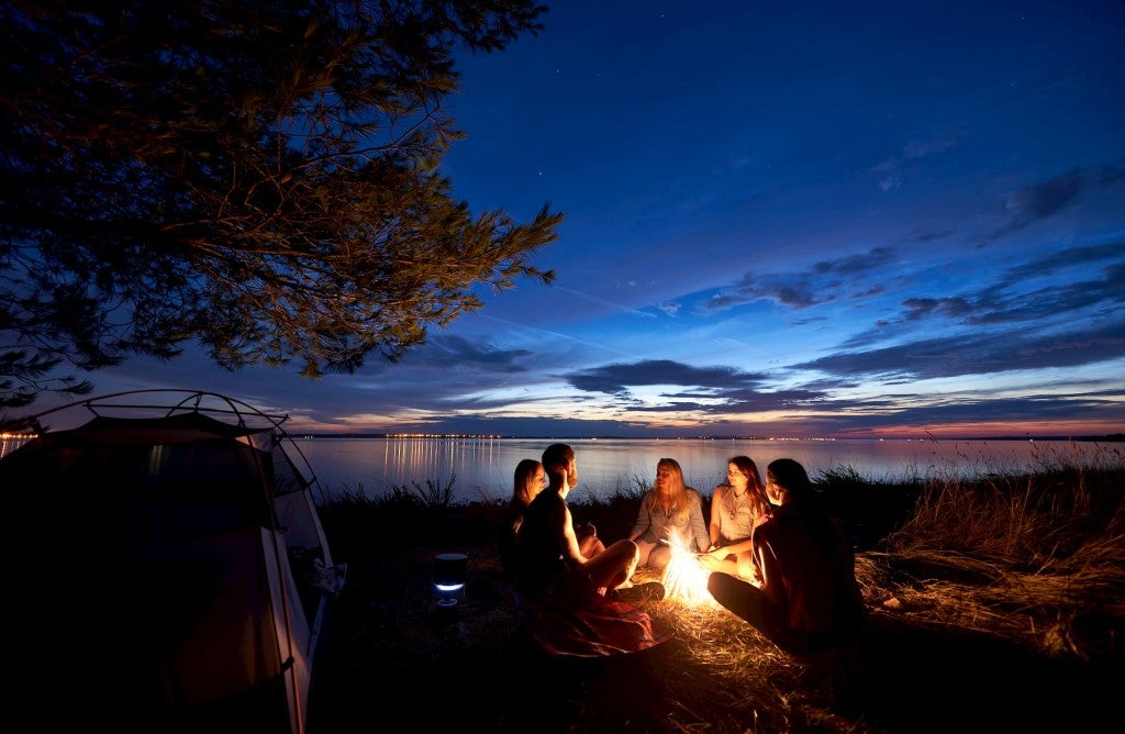 Night summer camping on sea shore. Group of five young tourists sitting on the beach around campfire near tent under beautiful blue evening sky.