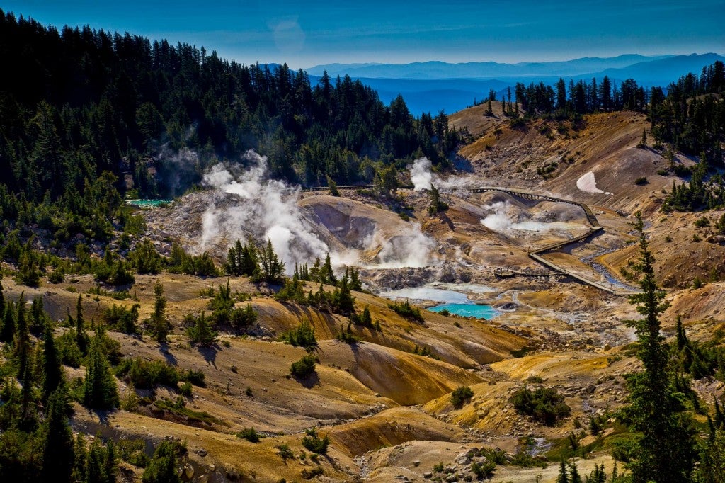 Textured geothermal valley with yellow tinted ground, bright blue ponds, winding path, and mountains in the background.
