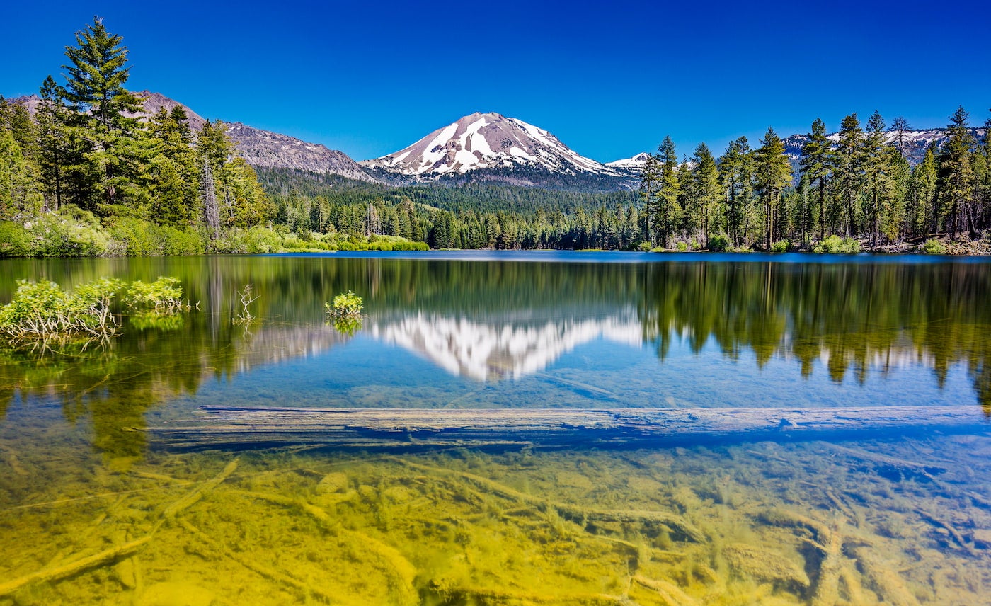 Mountain with snow patches reflects upon a lake in the foreground surrounded by an evergreen forest.