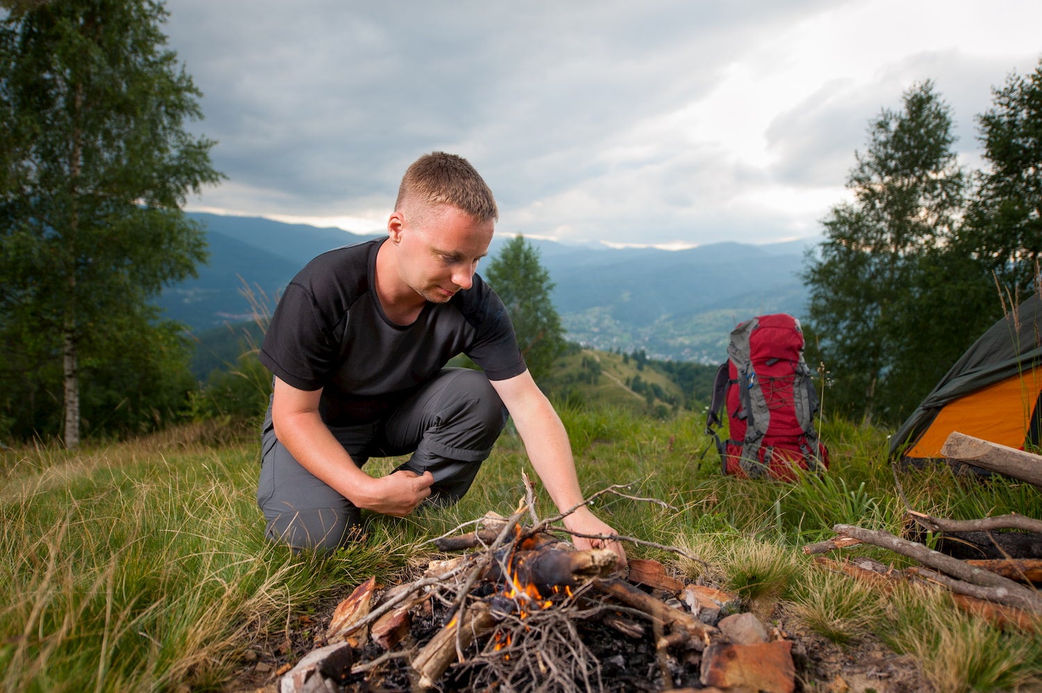 young man tourist kindling campfire on the hill with tent and travel backpack in background