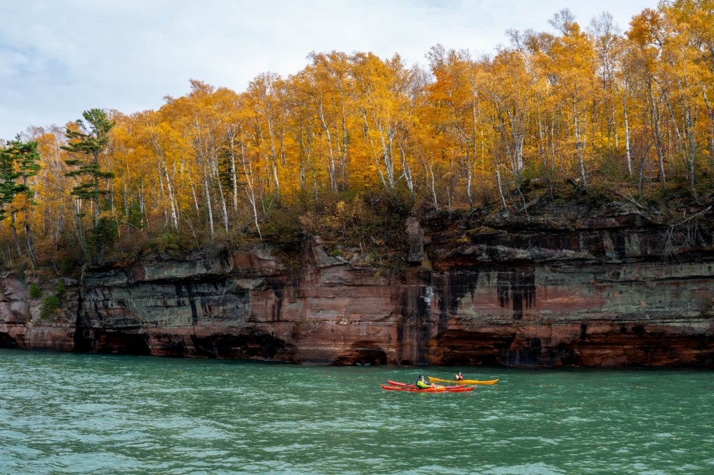 Two kayakers paddling beside a large forested cliff.