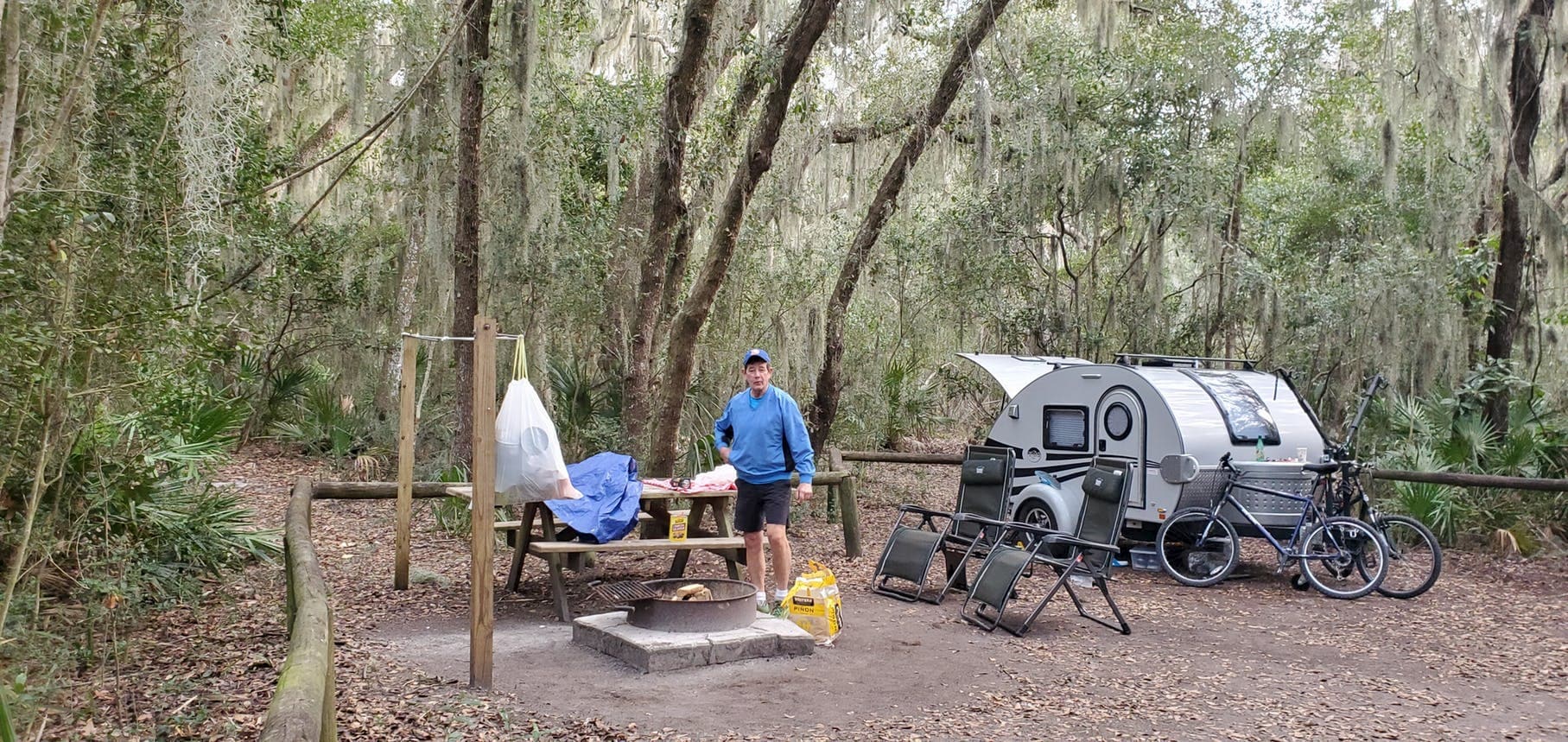 Man beside a teardrop trailer in mossy forested campsite.