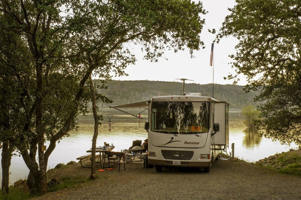 Camper parked beside water with people outside lounging in camp chairs.