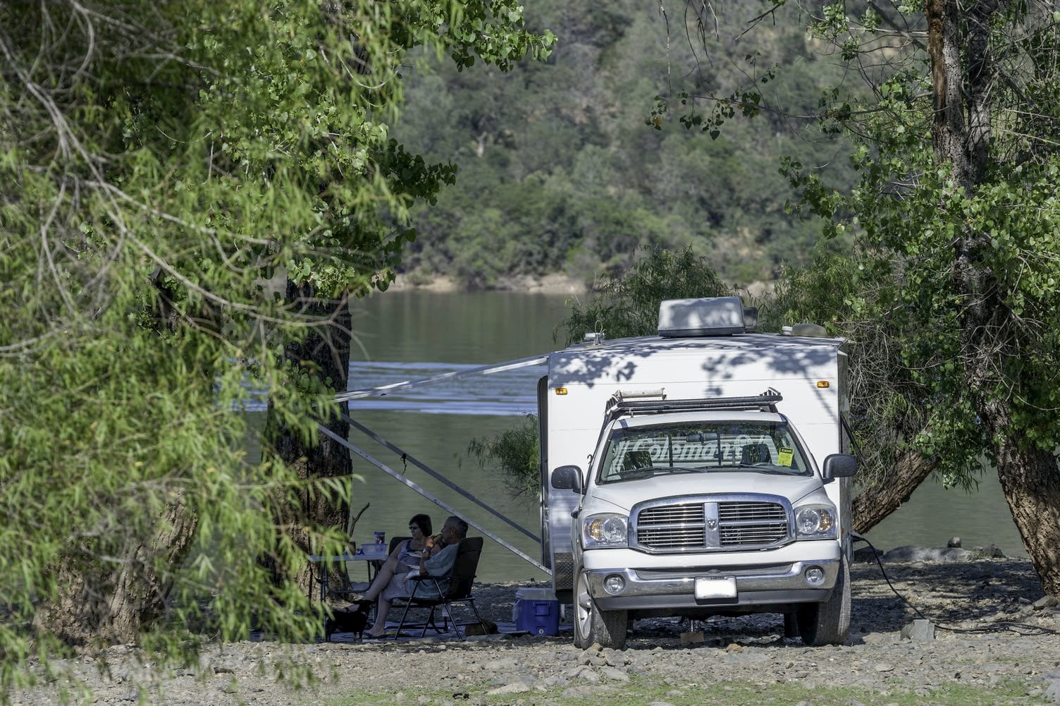 People lounging beside pickup truck with pull behind camper on a lake.
