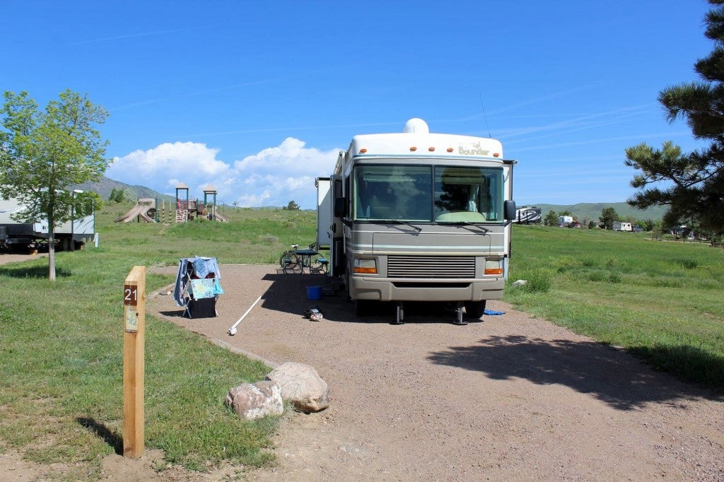 RV parked in gravel space beside lush green field on a clear sunny day.