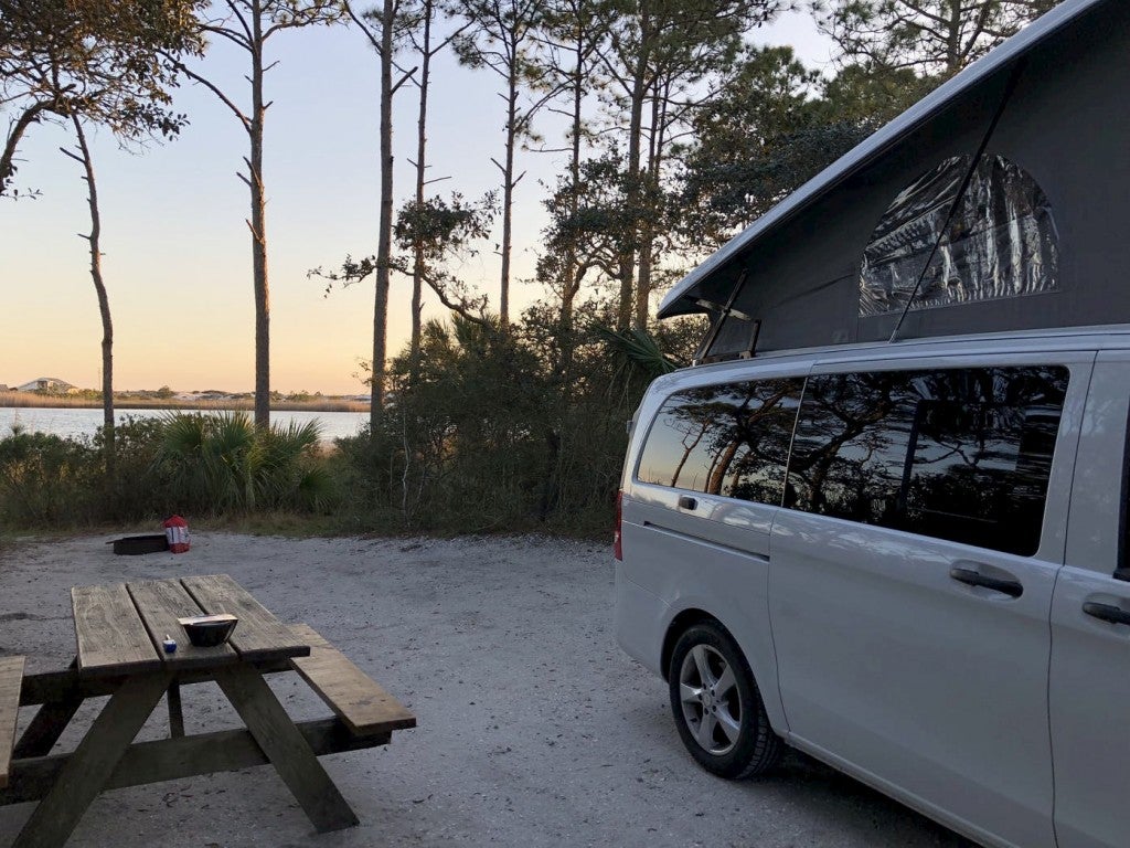 White van with pop top park beside picnic table and palm trees at coastal campsite.