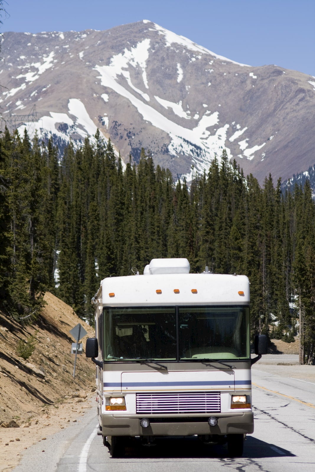 RV driving down highway with large mountains dotted with snow in the background.