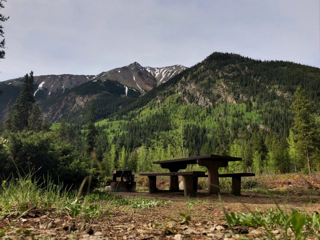 Picnic table and fire ring sit below epic mountain range with snow patches.