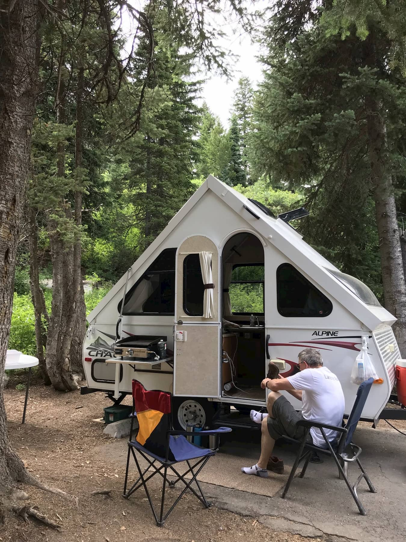 Man sitting in camping chair in front of A-line trailer in the forest.