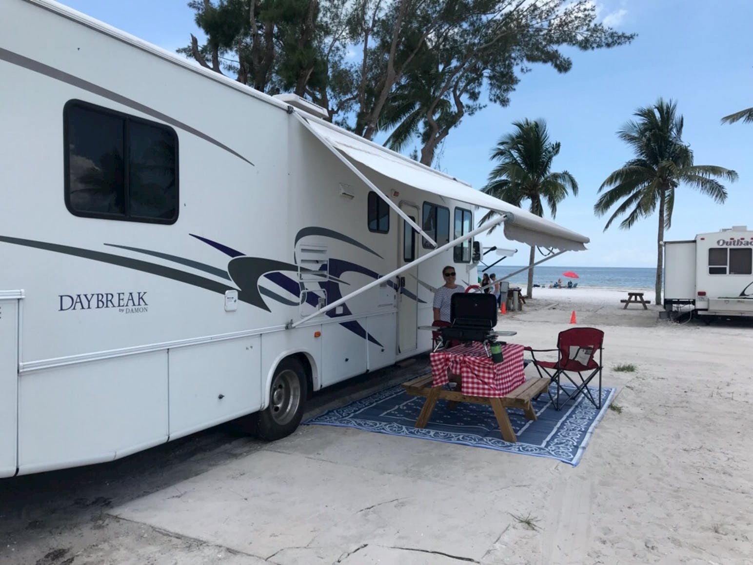 Women standing beside dining table and RV on the beach.