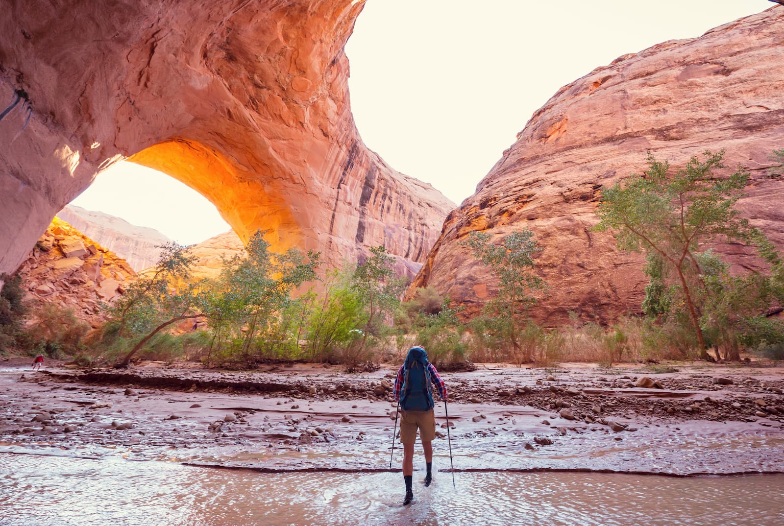 Biker with trekking poles wading through water below red rock arches.