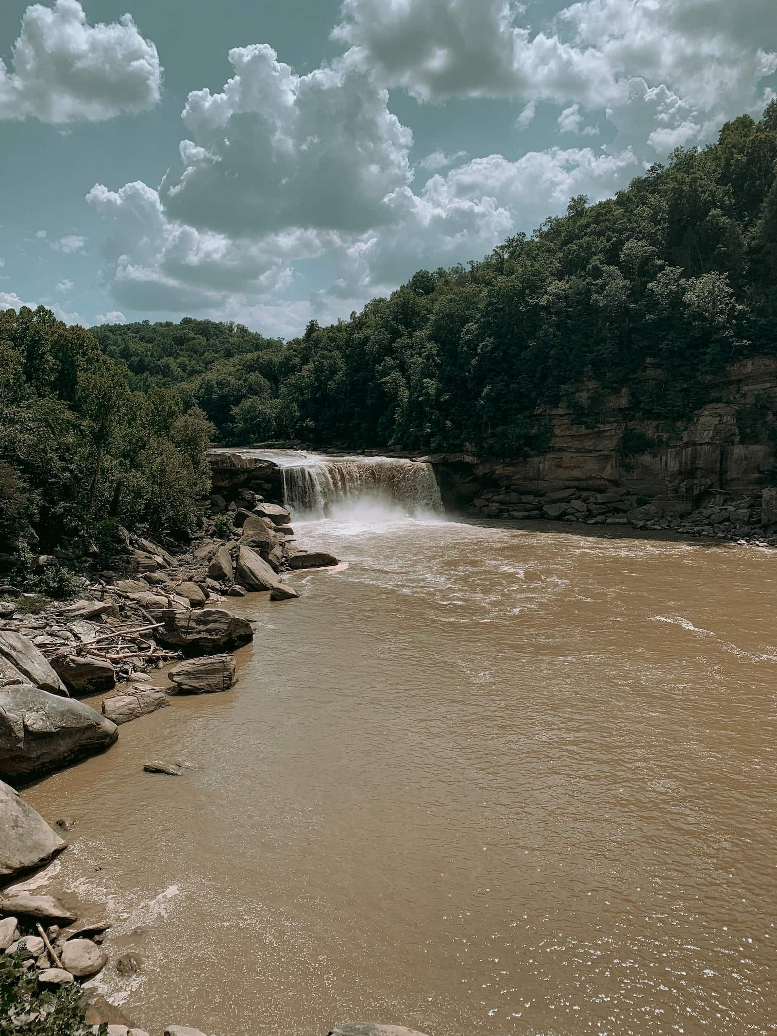 Waterfall pouring over muddy water under a cloudy bright blue sky.