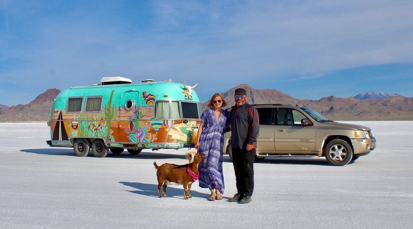 Couple poses in front of airstream with pick up truck and goat on salt flats.