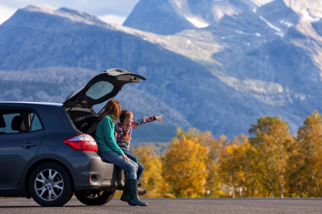 Young girl stand beside her mom in front of a car pointing out into an alpine landscape.