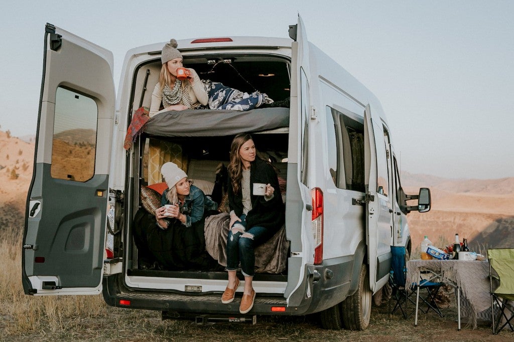 Three women drinking mugs of coffee in the back of a 2 level camper van rental.
