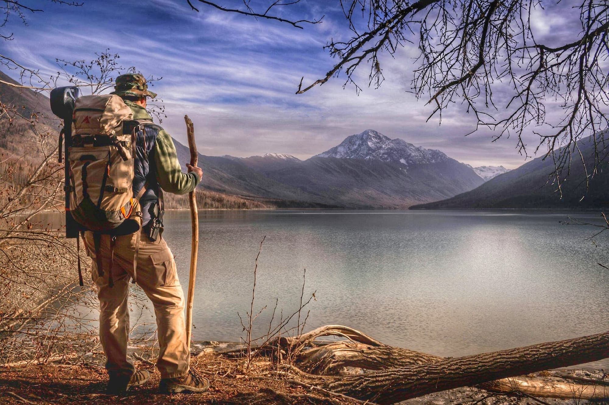 Hiker with camo backpack and wooden walking stick standing in fron of lake below a snowy mountain.