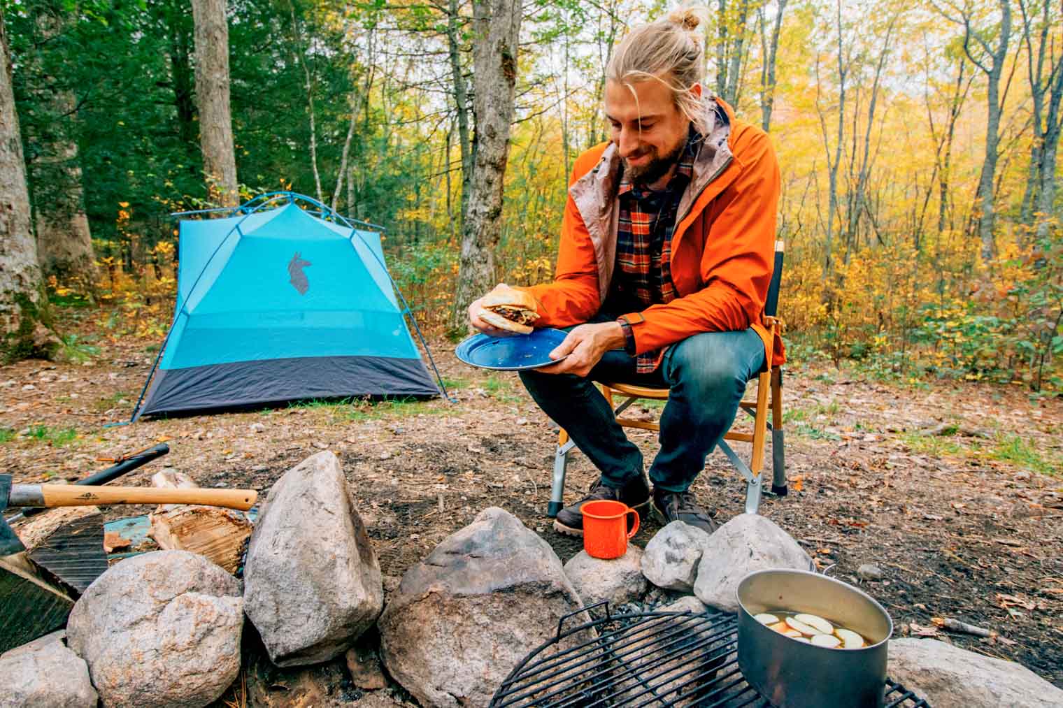 Man in orange jacket beside blue tent at a campsite enjoying an apple burger.