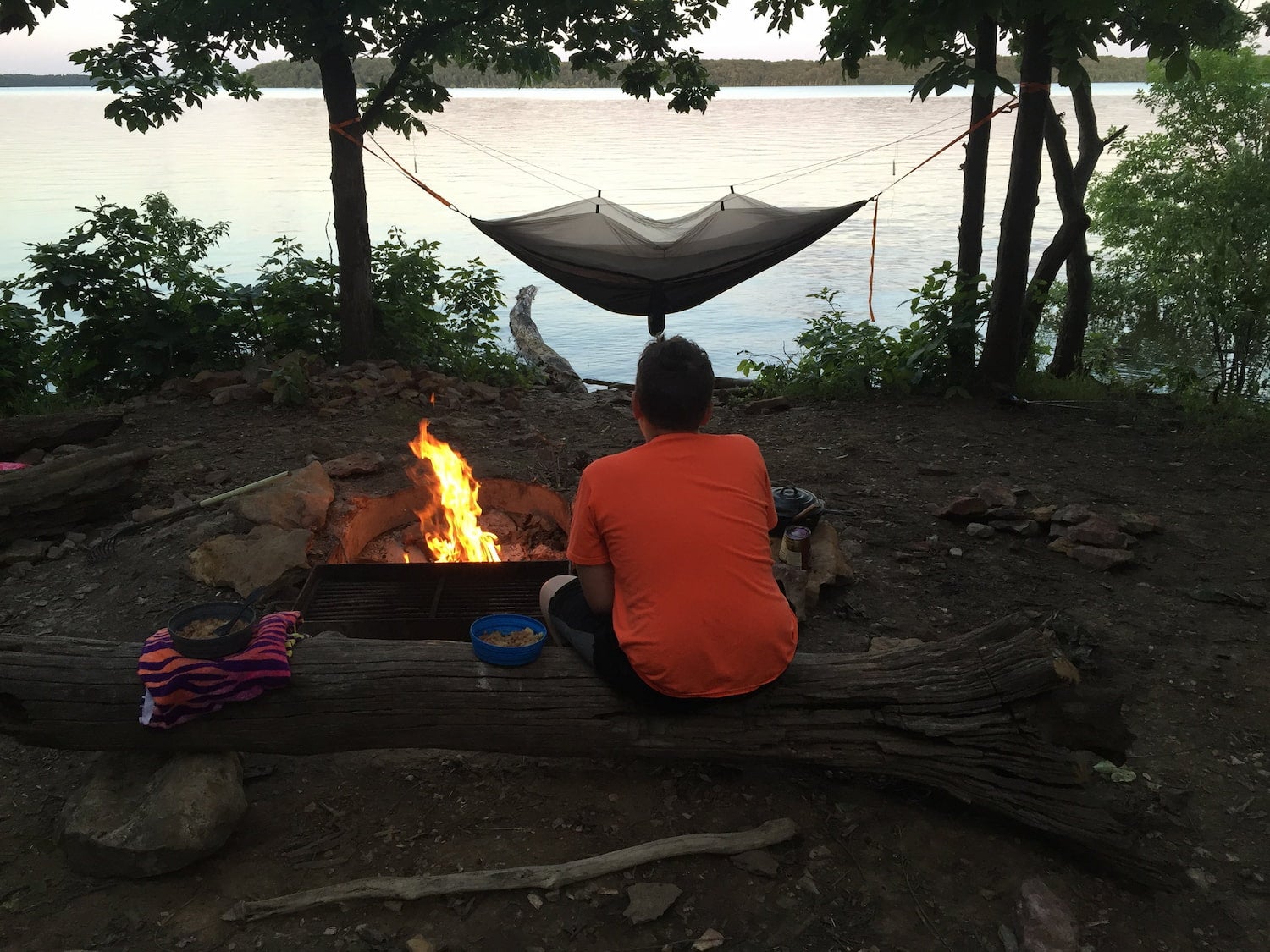 person sitting by fire with hammock and lake in background