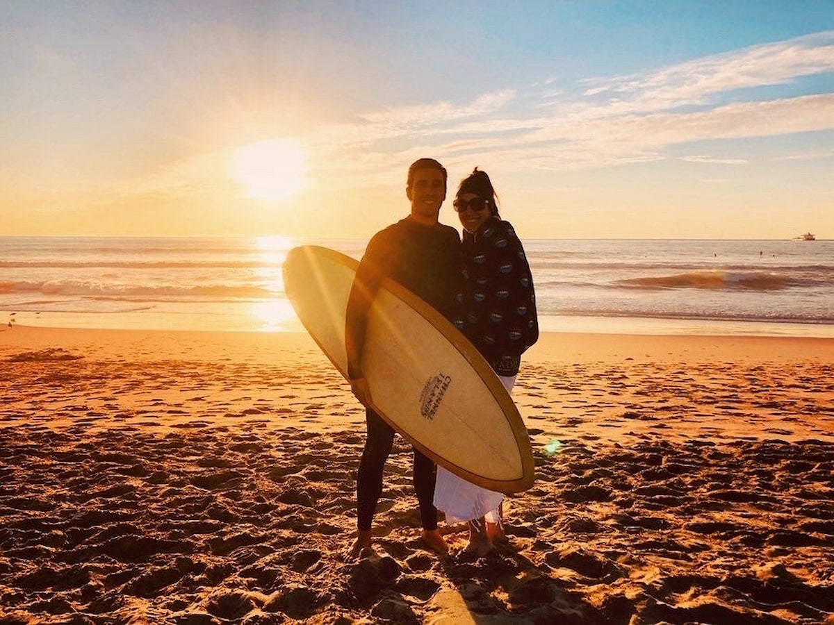 Couple posing on the beach at sunset with a surfboard.