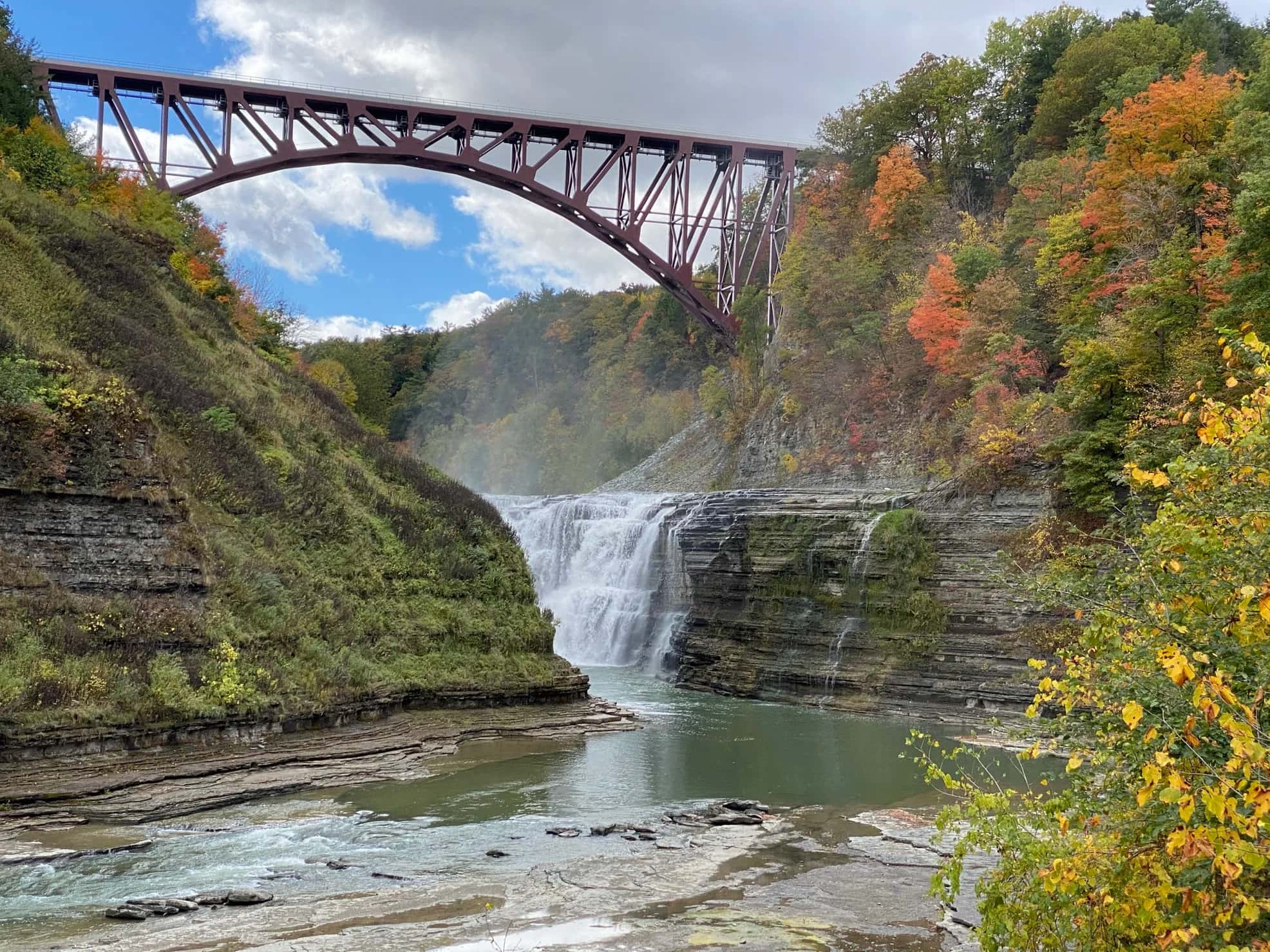 Iron bridge stretching over a rocky canyon with a waterfall down below surrounded by forests beginning to turn color in the autumn.
