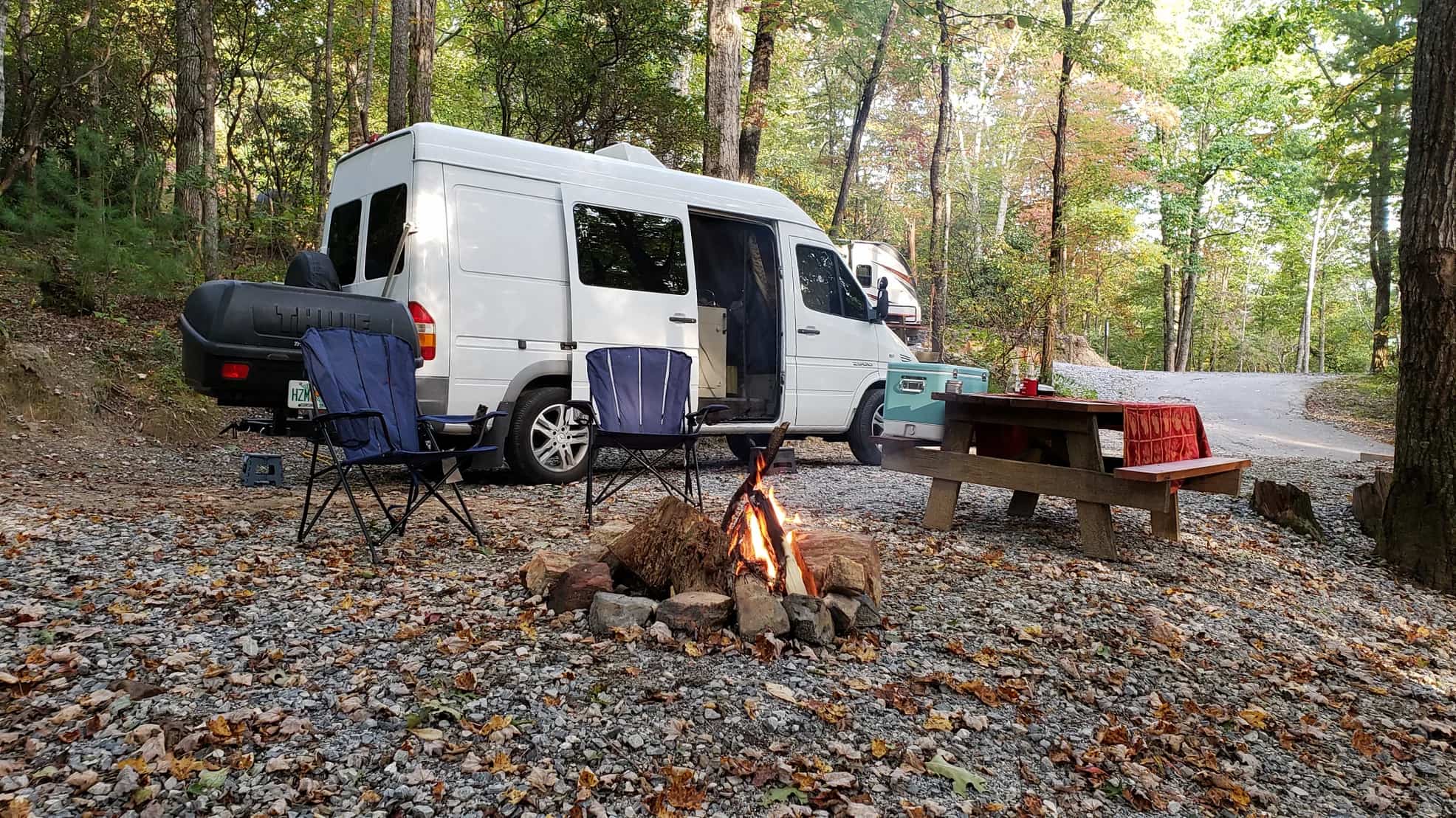 Van parked in campsite beside blazing campfire and picnic table.