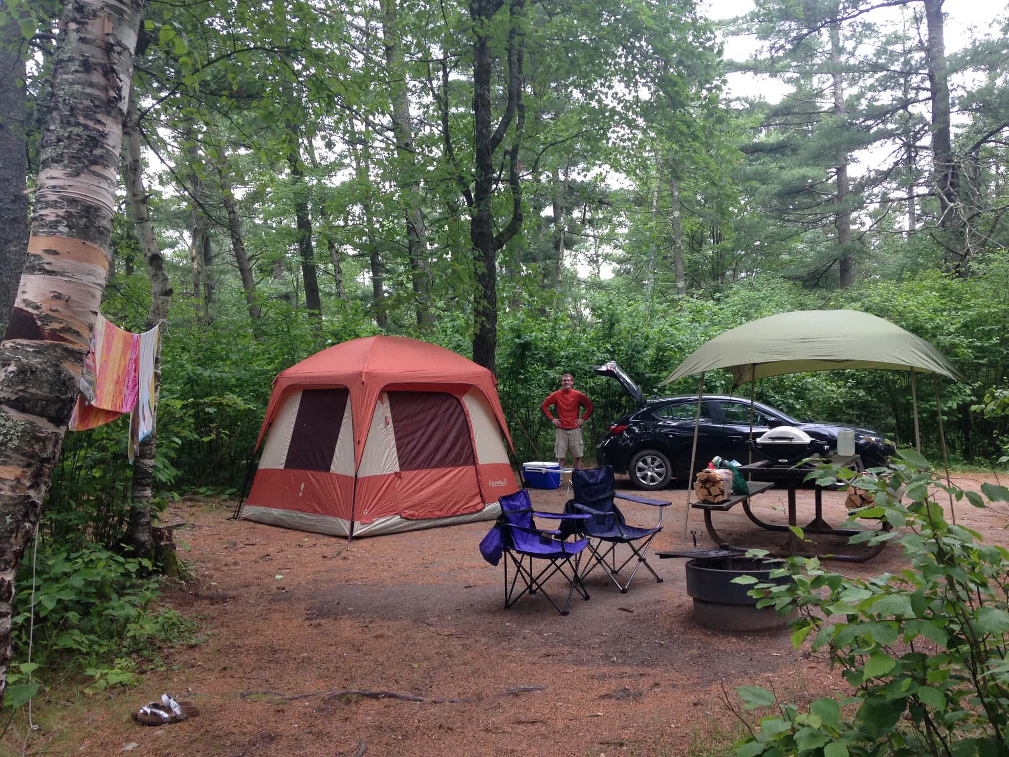 Campsite with red tent, awning, chairs and picnic table in a forested site.