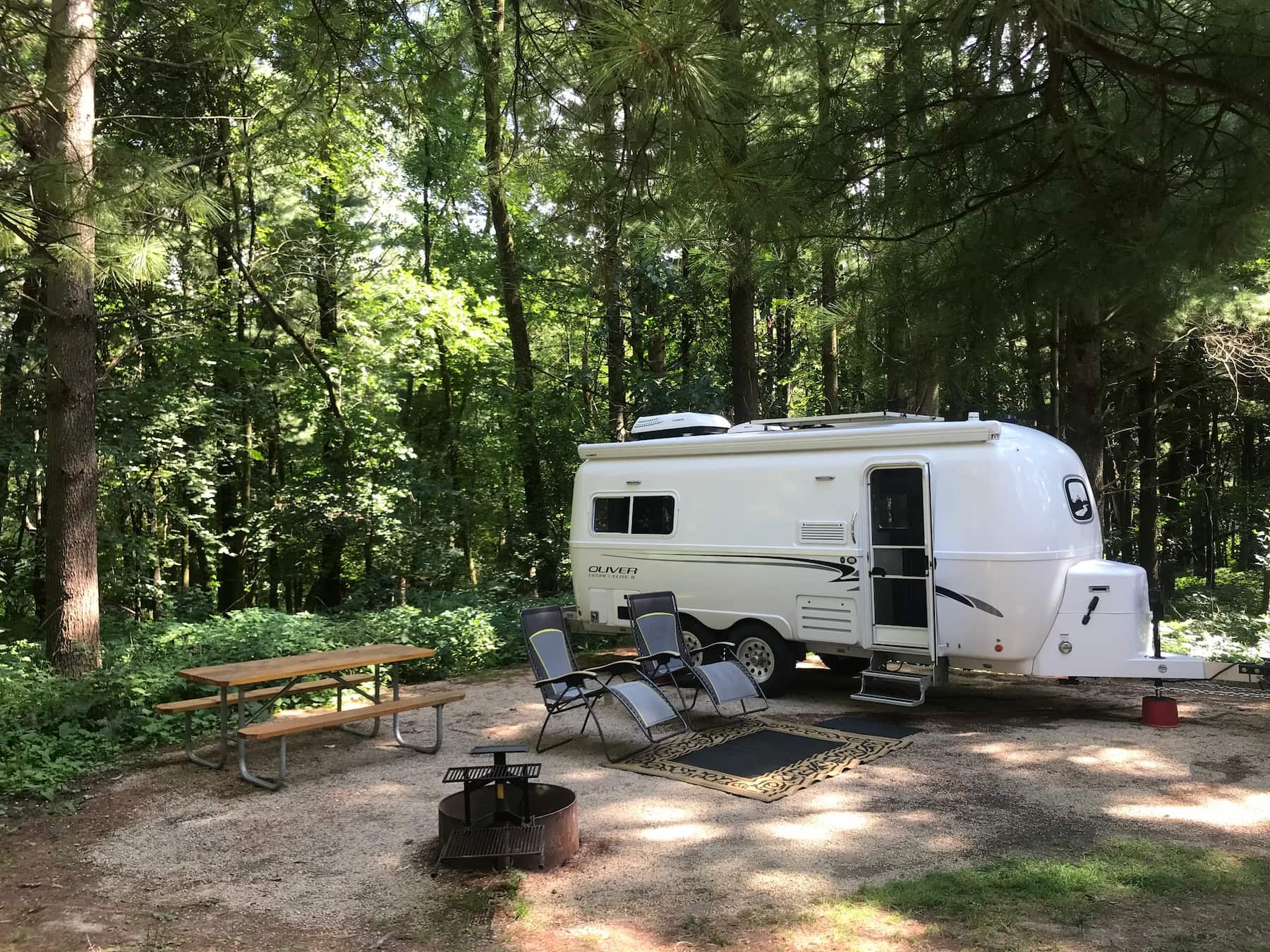 White trailer parked at a campsite beside camp chairs next to a picnic table in the forest.