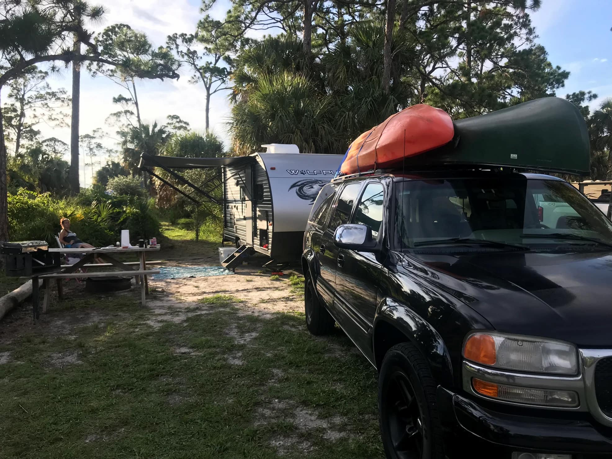 Truck with kayaks on top and trailer behind parked at a forested campsite on the beach in Florida.